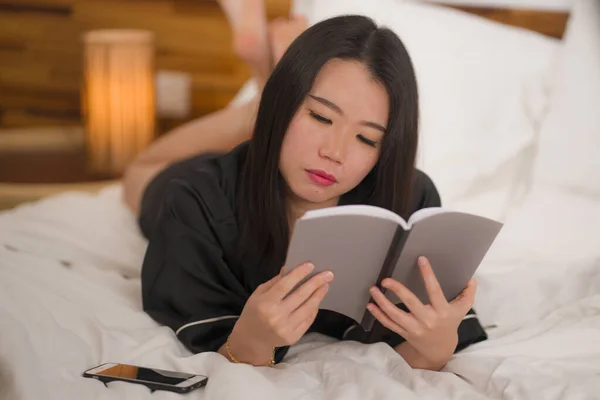 young beautiful and sweet Asian Chinese woman in bed reading book novel feeling happy and relaxed or studying at home in cute nightgown looking cozy and homey