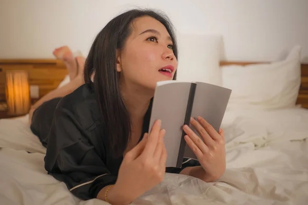 young beautiful and sweet Asian Chinese woman in bed reading book novel feeling happy and relaxed or studying at home in cute nightgown looking cozy and homey