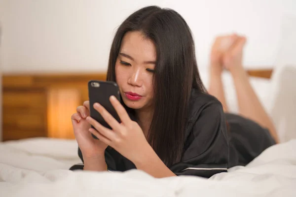 사랑 스러운 잠옷을 입고 침대에서 장난기있게 놀고 있는 젊고 아름다운 동양인 여성 이 소셜 미디어나 온라인 데이트 앱을 즐겁고 편안하게 즐기고 있다. — 스톡 사진