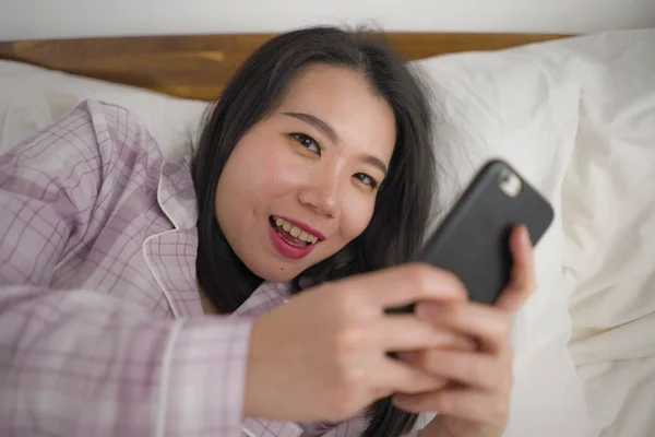 귀엽고 귀여운 잠옷을 입고 침대에서 장난기있게 놀고 있는 젊고 아름다운 동양인 여성 이 핸드폰을 사용하여 소셜 미디어나 온라인 데이트 앱을 즐겁고 편안하게 즐길 수있다. — 스톡 사진