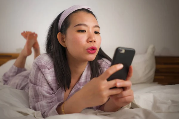 귀엽고 귀여운 잠옷을 입고 침대에서 장난기있게 놀고 있는 젊고 아름다운 동양인 여성 이 핸드폰을 사용하여 소셜 미디어나 온라인 데이트 앱을 즐겁고 편안하게 즐길 수있다. — 스톡 사진
