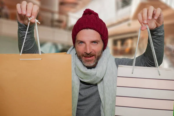 Młody szczęśliwy i atrakcyjny hipster człowiek w czapce zimowej i szalik uśmiechnięty trzymając torby na zakupy kupując prezenty świąteczne i prezenty podekscytowany i wesoły w centrum handlowym — Zdjęcie stockowe