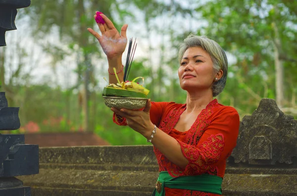 Bela e feliz mulher indonésia asiática vestida com tradicional costume religioso balinês segurando pau de incenso e flores oferecendo ao ar livre no templo de Bali — Fotografia de Stock