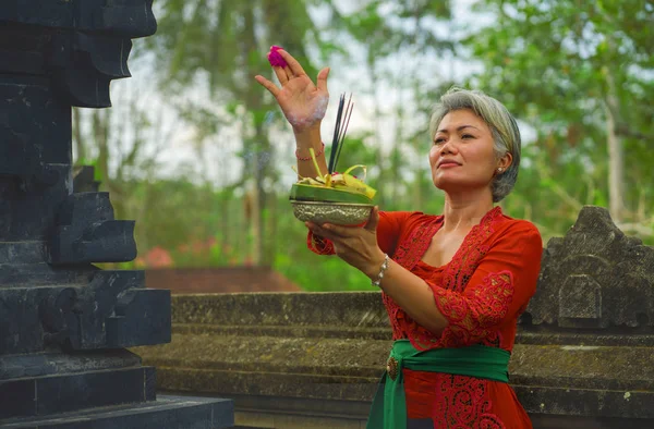 Bela e feliz mulher indonésia asiática vestida com tradicional costume religioso balinês segurando pau de incenso e flores oferecendo ao ar livre no templo de Bali — Fotografia de Stock