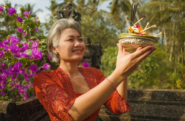 Bela e feliz mulher indonésia asiática vestida com tradicional costume religioso balinês segurando pau de incenso e flores oferecendo ao ar livre no templo de Bali — Fotografia de Stock