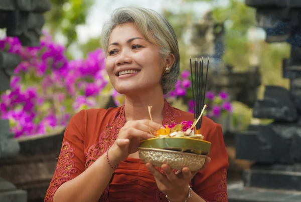 Bela e feliz mulher indonésia asiática vestida com tradicional costume religioso balinês segurando pau de incenso e flores oferecendo ao ar livre no templo de Bali — Fotografia de Stock