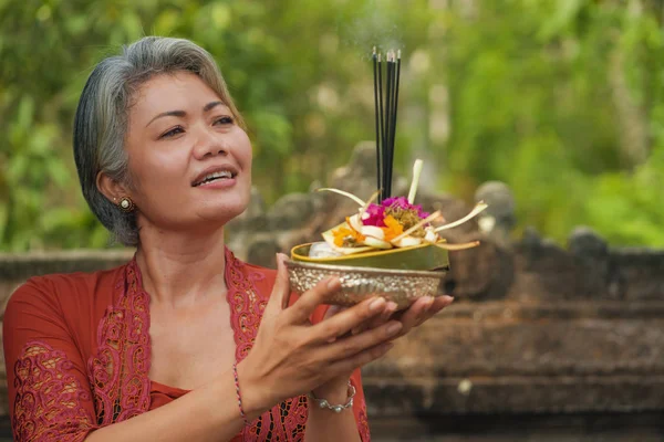 Gyönyörű és boldog ázsiai indonéz nő öltözött hagyományos balinéz vallási szokás gazdaság tömjén pálca és virágok kínál a szabadban Bali templom — Stock Fotó