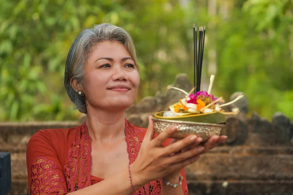 Bela e feliz mulher indonésia asiática vestida com tradicional costume religioso balinês segurando pau de incenso e flores oferecendo ao ar livre no templo de Bali — Fotografia de Stock