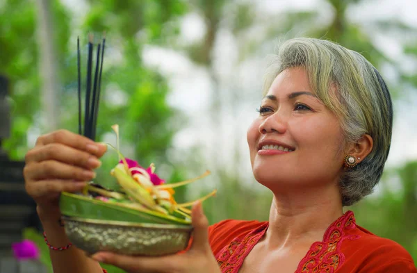 Bela e feliz mulher indonésia asiática vestida com tradicional costume religioso balinês segurando pau de incenso e flores oferecendo ao ar livre no templo de Bali — Fotografia de Stock