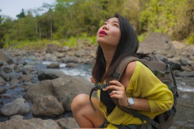 Genç, güzel ve mutlu Asyalı bir Japon kadın sırt çantasıyla dağları aşarak nehri geçiyor. Tatil doğasının tadını çıkarıyor ve neşeli bir yürüyüş yapıyor.