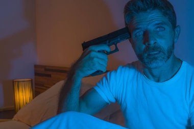 dramatic portrait in cinematic edgy lighting of young desperate and sick man pointing gun to his head for committing suicide shooting himself sitting on bed at night in horror 
