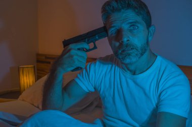 dramatic portrait in cinematic edgy lighting of young desperate and sick man pointing gun to his head for committing suicide shooting himself sitting on bed at night in horror 