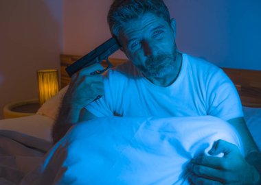 dramatic portrait in cinematic edgy lighting of young desperate and sick man pointing gun to his head for committing suicide shooting himself sitting on bed at night in horror 