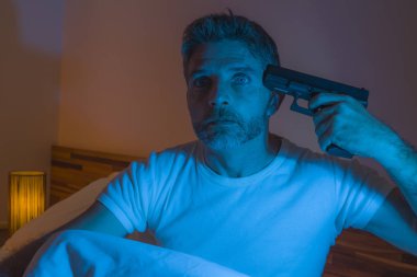 dramatic portrait in cinematic edgy lighting of young desperate and sick man pointing gun to his head for committing suicide shooting himself sitting on bed at night in horror 