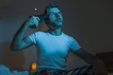 dramatic portrait in cinematic edgy lighting of young desperate and sick man pointing gun to his head for committing suicide shooting himself sitting on bed at night in horror 