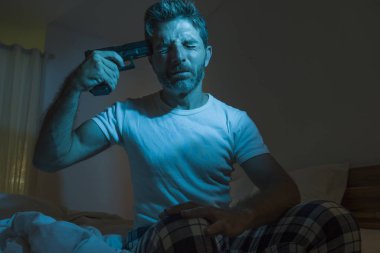 dramatic portrait in cinematic edgy lighting of young desperate and sick man pointing gun to his head for committing suicide shooting himself sitting on bed at night in horror 