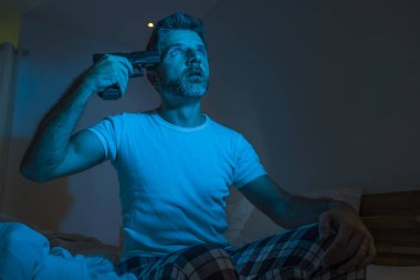 dramatic portrait in cinematic edgy lighting of young desperate and sick man pointing gun to his head for committing suicide shooting himself sitting on bed at night in horror 