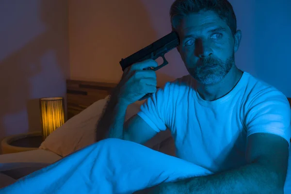 Dramatický portrét v kinematografickém ostrém osvětlení mladého zoufalého a nemocného muže mířícího pistolí na hlavu za spáchání sebevražedné střelby sedící v noci na posteli v hrůze — Stock fotografie