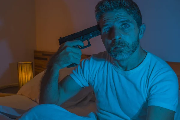 Potret dramatis dalam sinematik cahaya edgy dari pemuda putus asa dan orang sakit menunjuk pistol ke kepalanya untuk melakukan bunuh diri menembak dirinya duduk di tempat tidur di malam hari di horor — Stok Foto