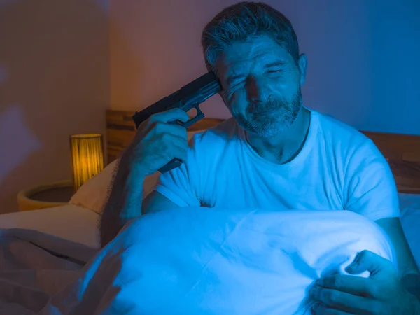 Dramatic portrait in cinematic edgy lighting of young desperate and sick man pointing gun to his head for committing suicide shooting himself sitting on bed at night in horror — ストック写真