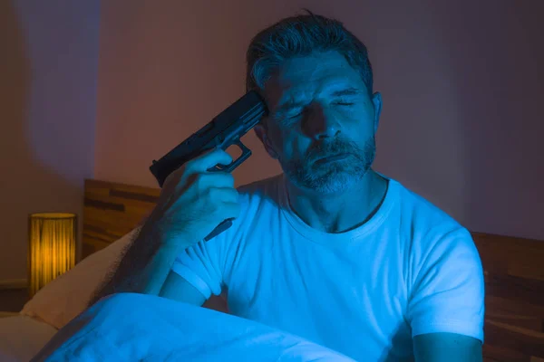 Dramatic portrait in cinematic edgy lighting of young desperate and sick man pointing gun to his head for committing suicide shooting himself sitting on bed at night in horror — ストック写真