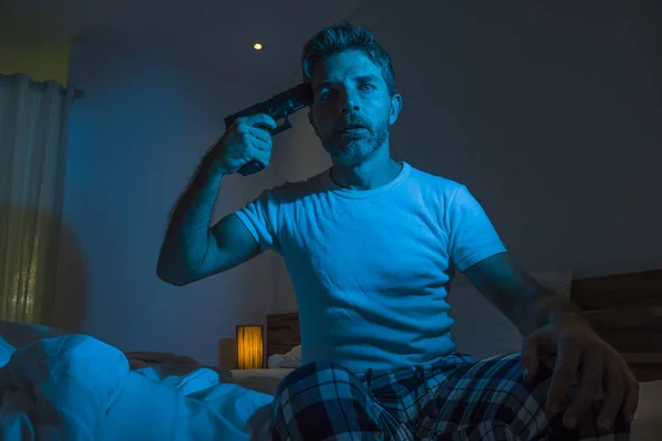 dramatic portrait in cinematic edgy lighting of young desperate and sick man pointing gun to his head for committing suicide shooting himself sitting on bed at night in horror