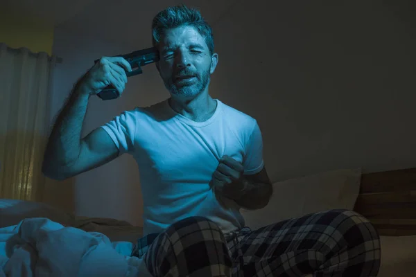 dramatic portrait in cinematic edgy lighting of young desperate and sick man pointing gun to his head for committing suicide shooting himself sitting on bed at night in horror