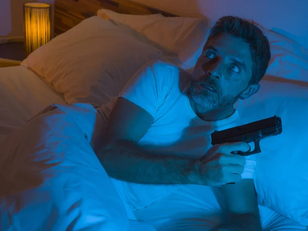 night edgy portrait of young stressed and paranoid American man lying on bed unable to sleep holding gun looking around scared feeling threatened suffering paranoia