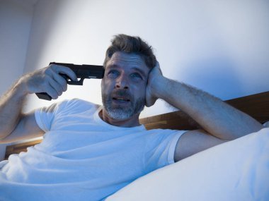 dramatic portrait in cinematic edgy lighting of young desperate and sick man pointing gun to his head for committing suicide shooting himself sitting on bed at night in horror 