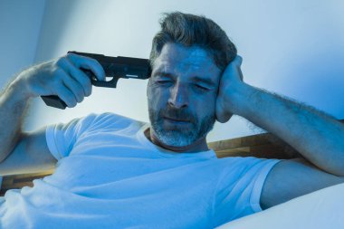 dramatic portrait in cinematic edgy lighting of young desperate and sick man pointing gun to his head for committing suicide shooting himself sitting on bed at night in horror 