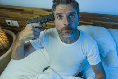 dramatic portrait in cinematic edgy lighting of young desperate and sick man pointing gun to his head for committing suicide shooting himself sitting on bed at night in horror 