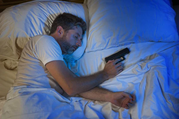 Retrato nocturno del joven estresado y paranoico hombre americano en la cama durmiendo y sosteniendo arma temiendo intruso irrumpiendo en casa en paranoia y concepto de autodefensa — Foto de Stock
