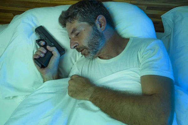 Éjszakai portré fiatal stresszes és paranoiás amerikai férfi az ágyban alszik, és a kezében fegyver félelmetes behatoló betör otthonába paranoia és önvédelem koncepció — Stock Fotó