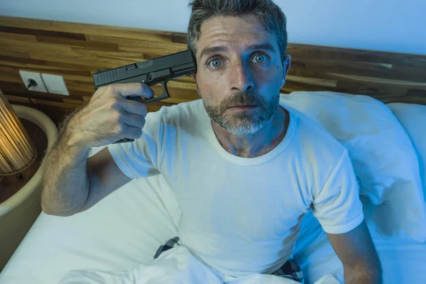 Dramatic portrait in cinematic edgy lighting of young desperate and sick man pointing gun to his head for committing suicide shooting himself sitting on bed at night in horror — Stock Photo, Image
