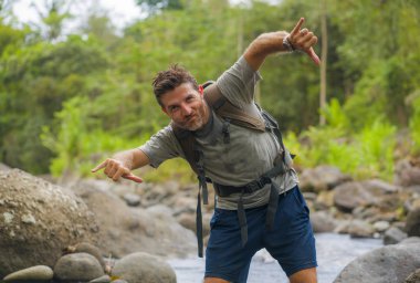 Genç, mutlu ve çekici bir adam seyahat çantasıyla ormanda yürüyüş yapıyor özgür hissediyor yaz yolculuğunda doğa ve çevrenin tadını çıkarıyor.