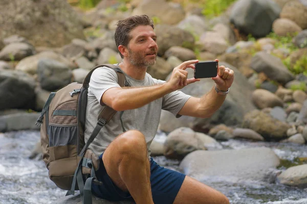 Ung glad och attraktiv man tar landskap foto med mobiltelefon innehav ryggsäck vandring i floden i skogen njuta av naturen bekymmersfri på sommaren vandring resa — Stockfoto