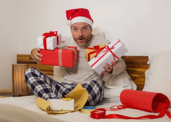 Imballaggio regali di Natale! giovane uomo felice e attraente seduto sul letto in cappello di Babbo Natale avvolgendo e preparando regali di Natale e scatole con carta e nastro sorridente eccitato — Foto Stock