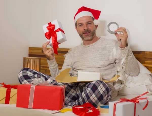 Noel hediyelerini paketliyorum! Genç, mutlu ve çekici bir adam Noel Baba şapkasıyla yatakta oturuyor. Noel hediyelerini ve kutuları kâğıt ve bant ile sarıp hazırlıyor. — Stok fotoğraf