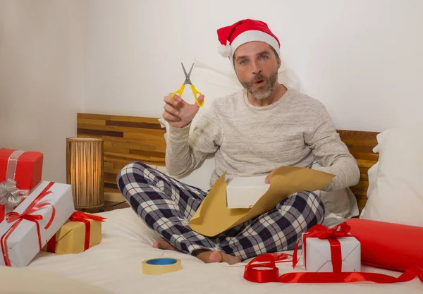 packing Chirstmas presents ! young happy and attractive man sitting on bed in Santa Claus hat wrapping and preparing xmas gifts and boxes with paper and tape smiling excited