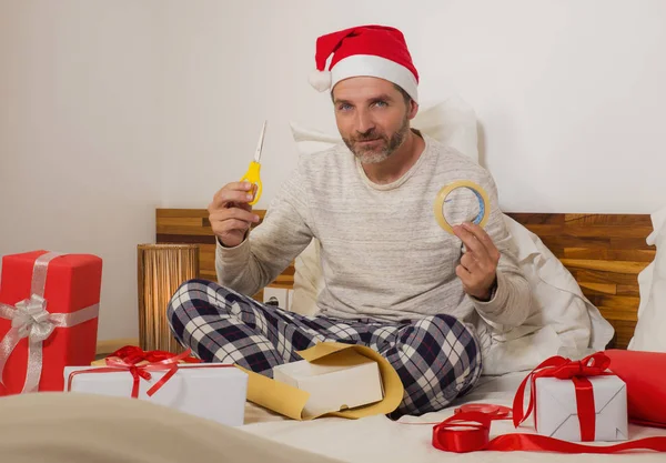 packing Chirstmas presents ! young happy and attractive man sitting on bed in Santa Claus hat wrapping and preparing xmas gifts and boxes with paper and tape smiling excited