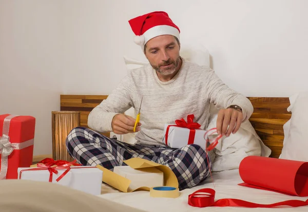 Imballaggio regali di Natale! giovane uomo felice e attraente seduto sul letto in cappello di Babbo Natale avvolgendo e preparando regali di Natale e scatole con carta e nastro sorridente eccitato — Foto Stock