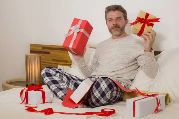packing Christmas presents ! young happy and attractive man sitting on bed cozy wrapping and preparing xmas gifts and boxes with paper and tape smiling excited