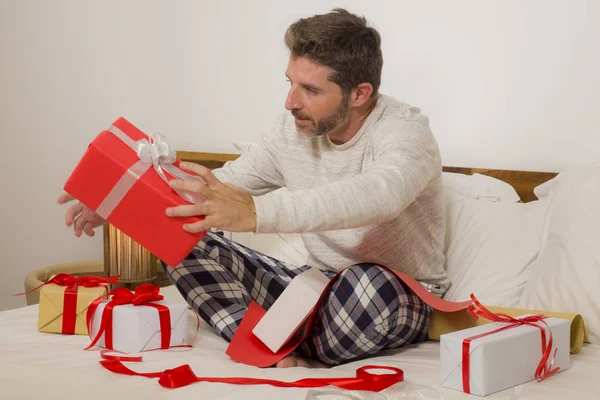 Imballaggio regali di Natale! giovane uomo felice e attraente seduto sul letto avvolgente accogliente e preparare regali di Natale e scatole con carta e nastro sorridente eccitato — Foto Stock