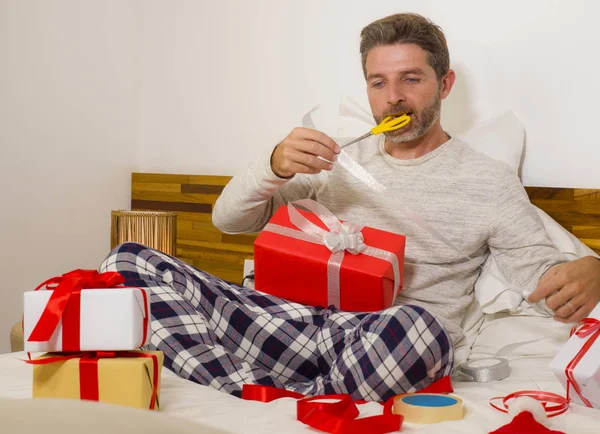 Imballaggio regali di Natale! giovane uomo felice e attraente seduto sul letto avvolgente accogliente e preparare regali di Natale e scatole con carta e nastro sorridente eccitato — Foto Stock