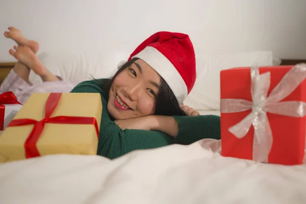 年轻美丽快乐的亚裔韩国女人头戴圣诞帽躺在床上，手里拿着许多圣诞礼物和礼品盒，兴奋而快乐地享受着圣诞生活 — 图库照片