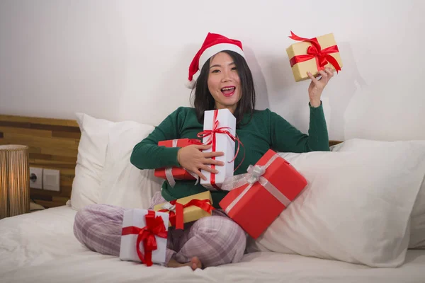 Χριστουγεννιάτικο πορτρέτο του τρόπου ζωής των νέων όμορφη και ευτυχισμένη ασιατική αμερικανική γυναίκα στο κρεβάτι στο καπέλο Άγιος Βασίλης κρατώντας πολλά δώρα Χριστούγεννα και κουτιά δώρων ενθουσιασμένοι και χαρούμενα — Φωτογραφία Αρχείου