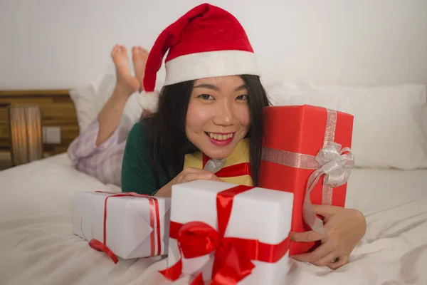 年轻美丽快乐的亚裔美国女人头戴圣诞帽躺在床上，拿着许多圣诞礼物和礼品盒的圣诞生活方式画像，兴奋而快乐 — 图库照片