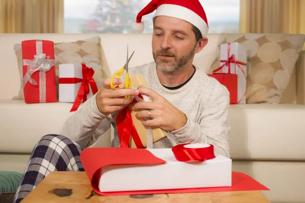 Natale vacanza stile di vita ritratto di giovane uomo attraente e felice in cappello di Babbo Natale a casa preparare regali di Natale confezionamento regali e scatole con carta e nastro — Foto Stock