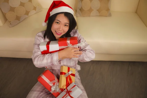 Χριστουγεννιάτικο πορτρέτο τρόπο ζωής διακοπών στο σπίτι των νέων όμορφη και ευτυχισμένη ασιατική αμερικανική γυναίκα με πιτζάμες και Σάντα καπέλο κρατώντας πολλά δώρα και κουτιά δώρων χαμογελώντας — Φωτογραφία Αρχείου