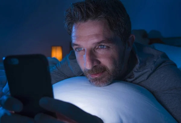 Hombre feliz con ojos azules acostado en la cama tarde en la noche en la red de luz oscura en el teléfono móvil o en línea citas sonriente relajado disfrutando de Internet en la adicción a las redes sociales —  Fotos de Stock